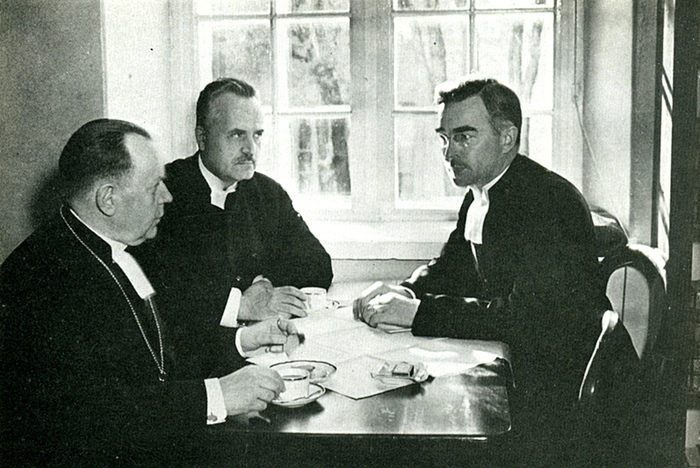 Svartvit bild där tre män i kaftan sitter vid ett bord och samtalar.