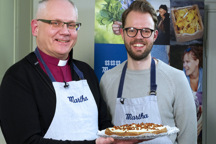 Biskop Åstran står bredvid en man i glasögon och håller upp ett fat med en tårta, båda har vita förkläden med texten Martha i blått.